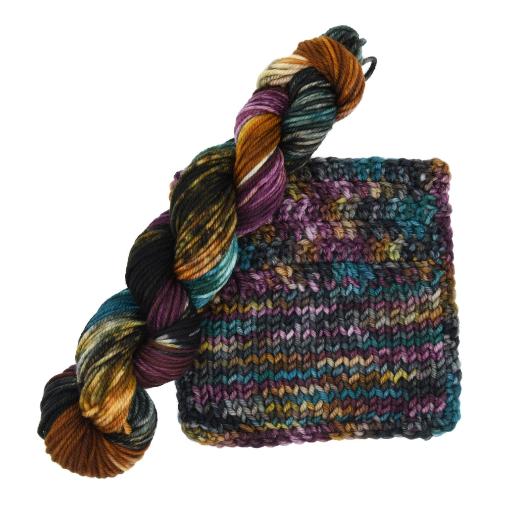 Skein and knit and crochet sample of Crown & Cloak hand-dyed yarn by Woolly Headed Ideas featuring a moody blend of black, dark gray, teal, green, olive, and a splash of gold.