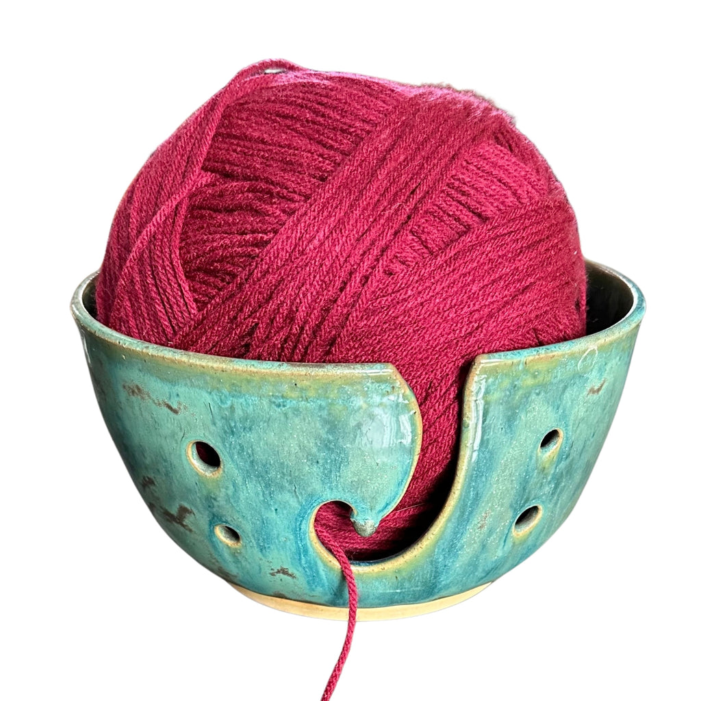 A ball of deep magenta yarn resting inside a turquoise glazed ceramic knitting bowl, shown on a plain white background.
